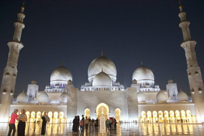 a large white building with domes and lights with Sheikh Zayed Mosque in the background
