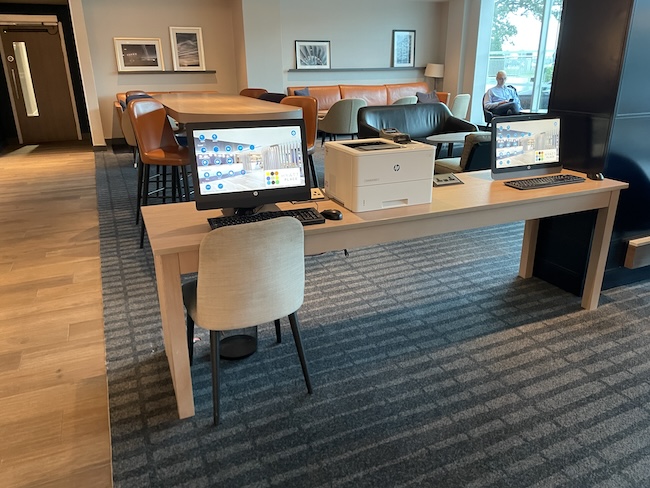 a desk with computers and chairs in a room with a man sitting in the background